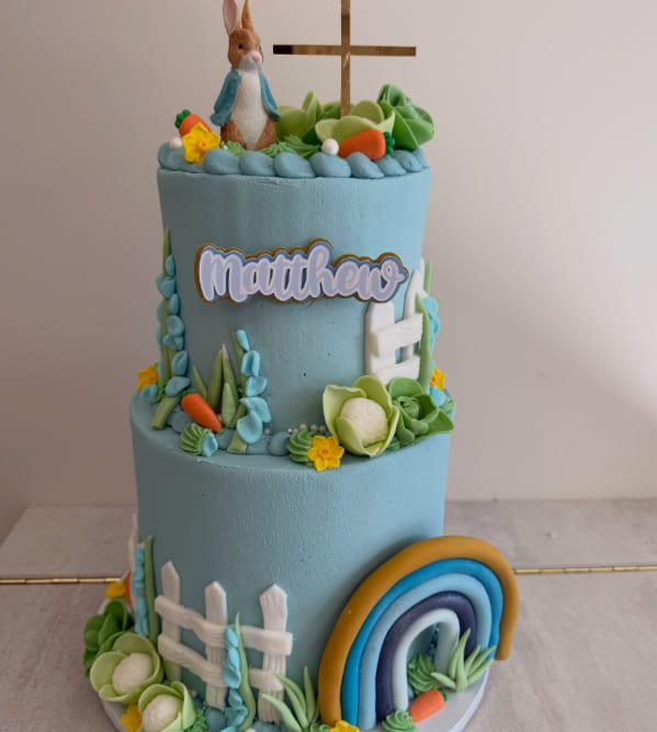 A two-tier powder-blue christening cake with a Peter Rabbit figure, gold cross, fondant rainbow and "Matthew" plaque