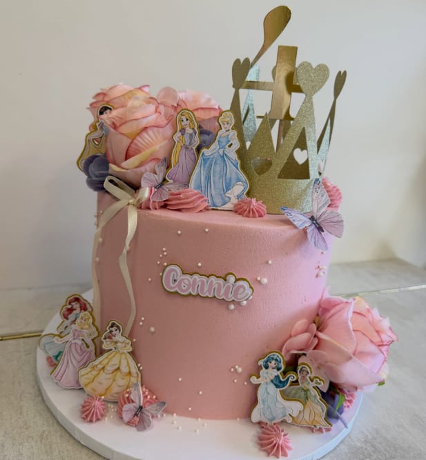 A candy-pink buttercream birthday cake with a gold crown topper, Disney princess figures and a "Connie" plaque