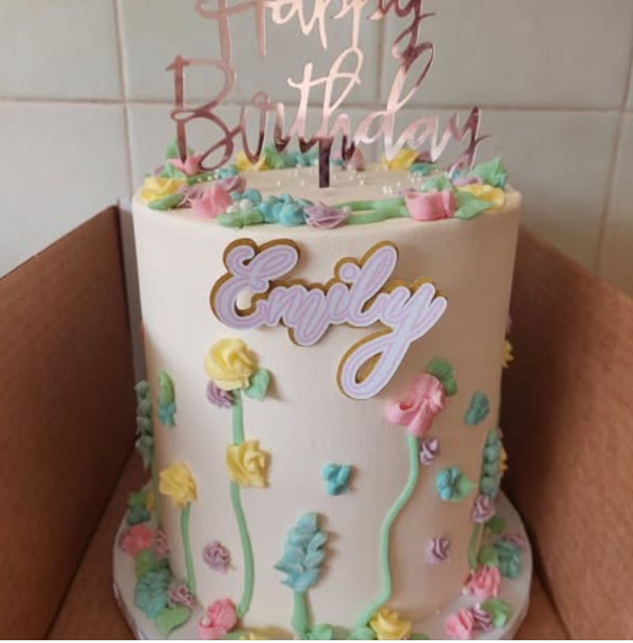 A tall cream cake hand-piped with pastel buttercream wildflowers and a rose-gold Happy Birthday topper