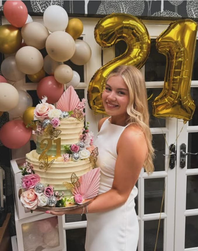 Poppy holding a two-tier 21st birthday cake decorated with roses, butterflies and disco balls, in front of gold '21' balloons